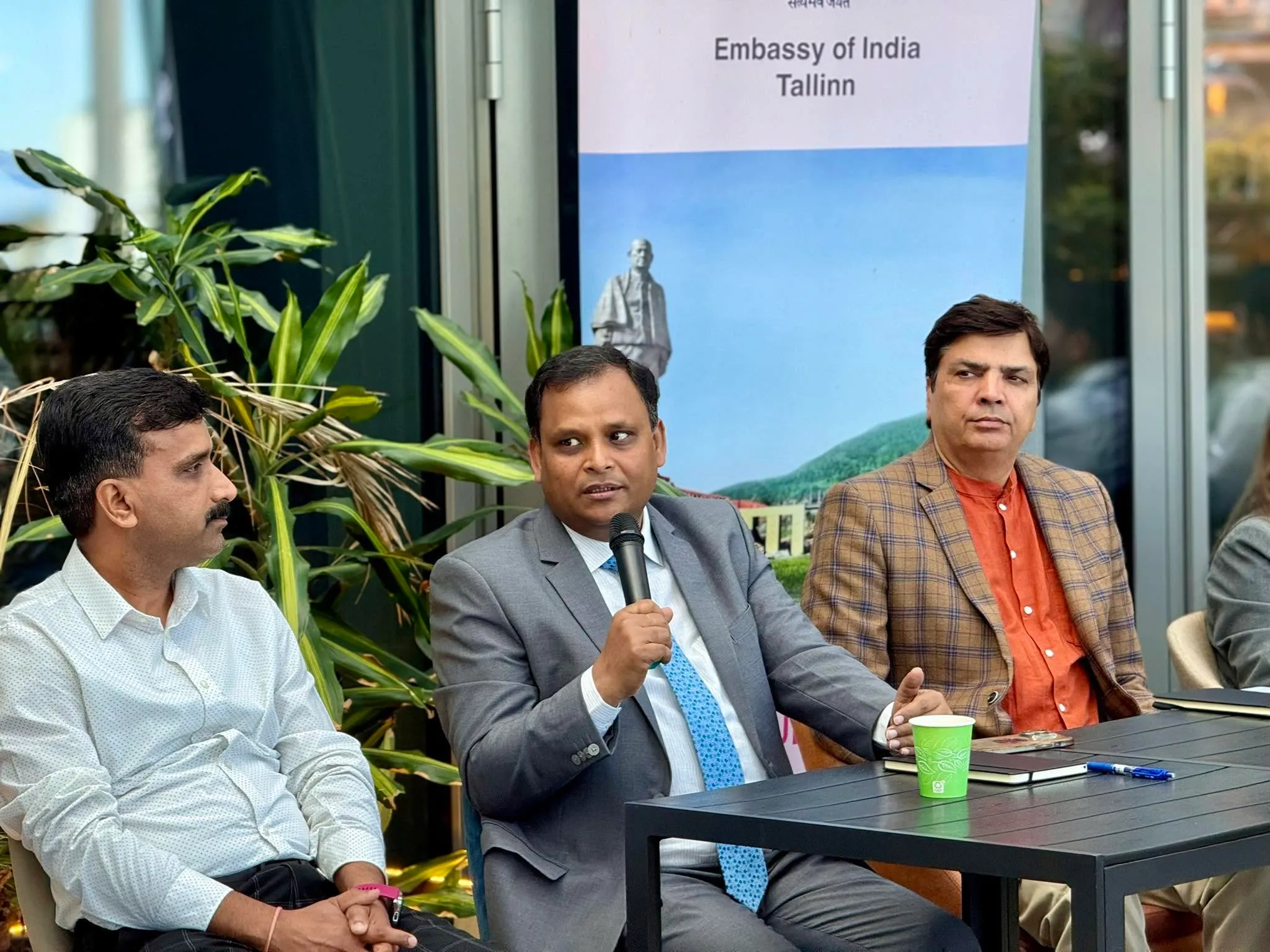 H.E. Ambassador Ashish Sinha speaking into a microphone at a table, seated between two guests, with an Embassy of India banner behind